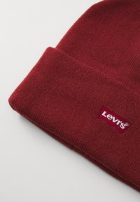 Rode gebreide beanie met een omgeslagen rand, voorzien van een klein wit Levi's logo patch. De textuur is zacht en licht geribbeld.