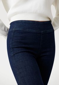 Jean en denim noir haut de taille avec une texture lisse, présentant une poche frontale et un détail de couture subtil à la taille.