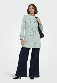 Femme portant un trench-coat vert clair ceinturé, un jean large foncé, des escarpins noirs et un sac à bandoulière noir.