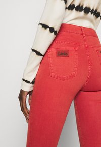 Czerwone dżinsy z wysokim stanem, wykonane z denimu, z kieszonką naszywaną z metką z logo. Modelka ma na sobie bluzkę z długim rękawem w stylu tie-dye.