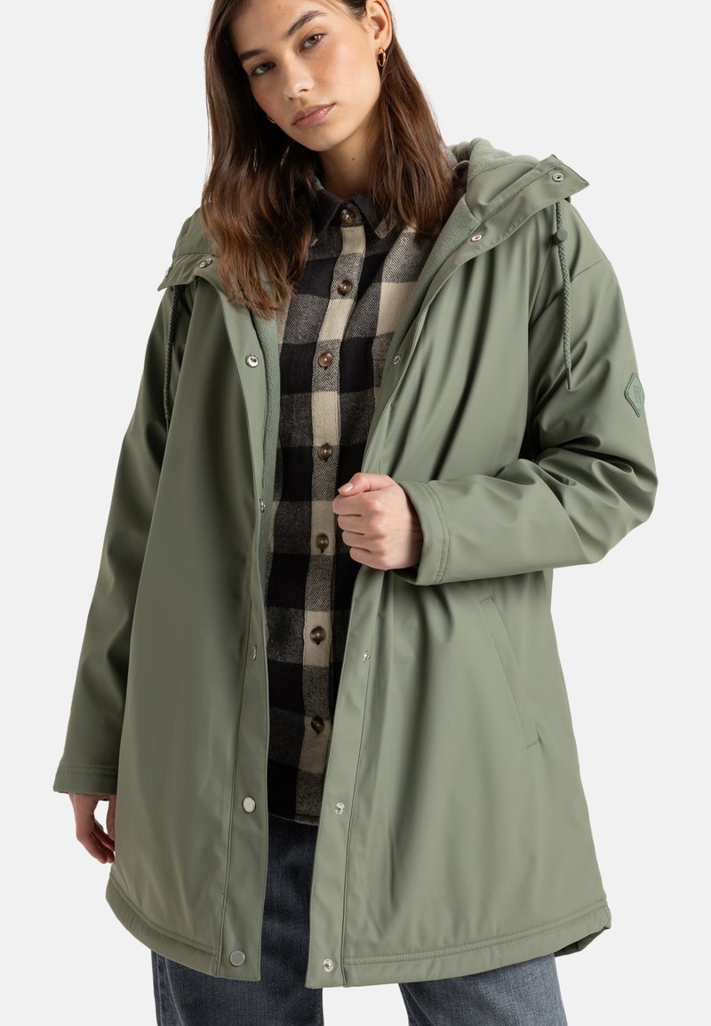 Olijfgroene waterdichte jas met een trekkoordcapuchon en drukknoopsluitingen, gedragen over een zwart-beige geruit overhemd, met zijzakken.