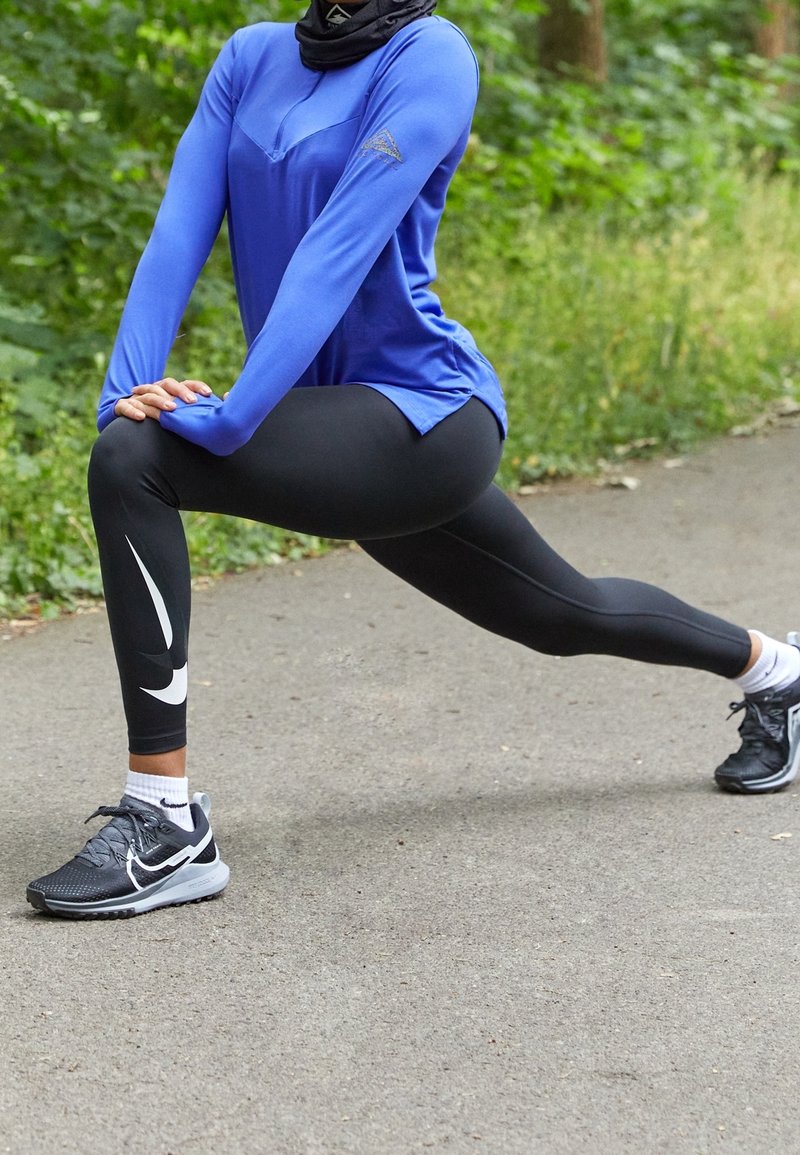 Haut de sport à manches longues bleu, leggings noirs avec logo Nike blanc, et chaussures de sport noires. Personne en position de fente sur un chemin.