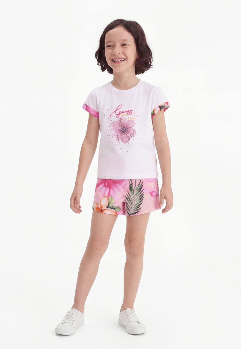 Χαμογελαστή κοπέλα με κοντά σκούρα μαλλιά, που φοράει λευκό μπλουζάκι με ροζ λουλουδάτη εκτύπωση και ροζ σορτς με λουλούδια, συνδυασμένα με λευκά αθλητικά παπούτσια.