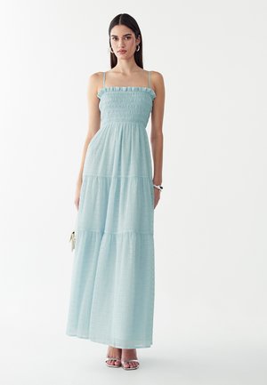 Mujer que viste un vestido largo azul claro, sin mangas, con capas, tirantes finos y escote con volantes, de pie frente a un fondo liso.