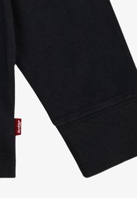 Crna pamučna košulja s dugim rukavima, s rebrastim manžetama i malom crvenom Levi's oznakom sa strane. Prikaz detalja rukava izbliza.