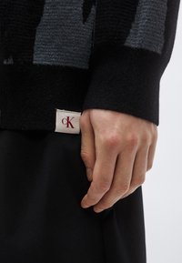 Črn pulover z sivim vzorčastim pletenjem, z rebrastim manšetom. Majhna bela etiketa z napisom "cK" je vidna blizu roba.