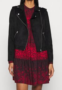 Giacca da moto in suede nera con zip argentate, colletto a falda ed epaulette, indossata sopra un abito floreale rosso con orlo a balze.