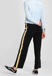 Svarta byxor med vida ben, med gula och vita sidoränder. Bärs med en ljusblå denimjacka och svarta sneakers.