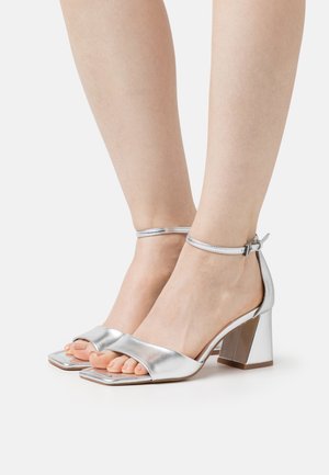 Sølv højhælede sandaler med firkantet tå, bred rem og justerbar ankelspænde. Tekstureret metalfinish og blokhæl.