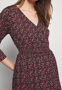 Schwarzes Blumenkleid mit kleinen roten und weißen Blumenmustern, V-Ausschnitt, geraffter Taille und Dreiviertelärmeln. Weicher Stoffhinweis.