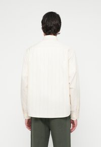 Sötét hajú férfi, aki hosszú ujjú krém színű inget és olívazöld nadrágot visel, háttal áll egy sima fehér háttér előtt.