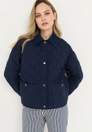 Junge Frau mit langen blonden Haaren trägt eine marineblaue gesteppte Jacke mit Messingknöpfen und schwarz-weiß gestreifte Hosen, steht vor einer weißen Wand.