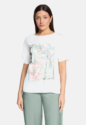 Femme portant un haut blanc à manches courtes avec un motif floral et de feuilles pastel, assorti à un pantalon large vert clair, debout devant un fond blanc.