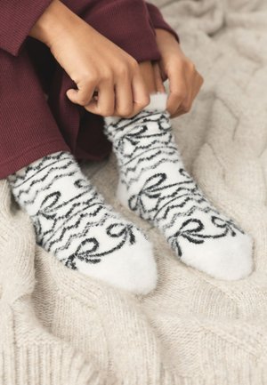 Hände ziehen an weißen, flauschigen Socken mit schwarzen Wirbelmustern, getragen von einer Person in burgunderfarbenen Hosen, die auf einer beige gestrickten Decke sitzt.
