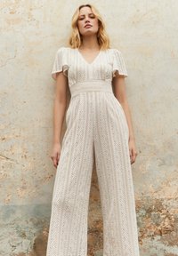 White lace jumpsuit with a V-neck, short flutter sleeves, and wide legs featuring a geometric pattern and textured waistband.