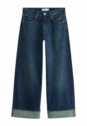 Kvinde står udendørs iført en hvid ærmeløs top og brede, opsmøgede mørke jeans med mønstrede sko mod en grøn og hvid væg.