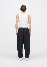 Vīrietis baltā bezroku krekliņā, melnās sporta biksēs ar aizmugurējo kabatu un zilā logotipu, kā arī baltās sporta kurpēs, stāvot pret baltu fonu.