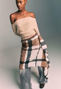 Beige off-the-shoulder sweater with ribbed hem, paired with a brown and white plaid wrap skirt. Model wears loose gray jeans and dark boots.