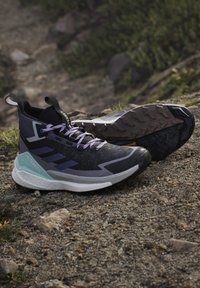 Paire de chaussures de trail noires et violettes avec des semelles épaisses reposant sur un terrain gravillonné en plein air.