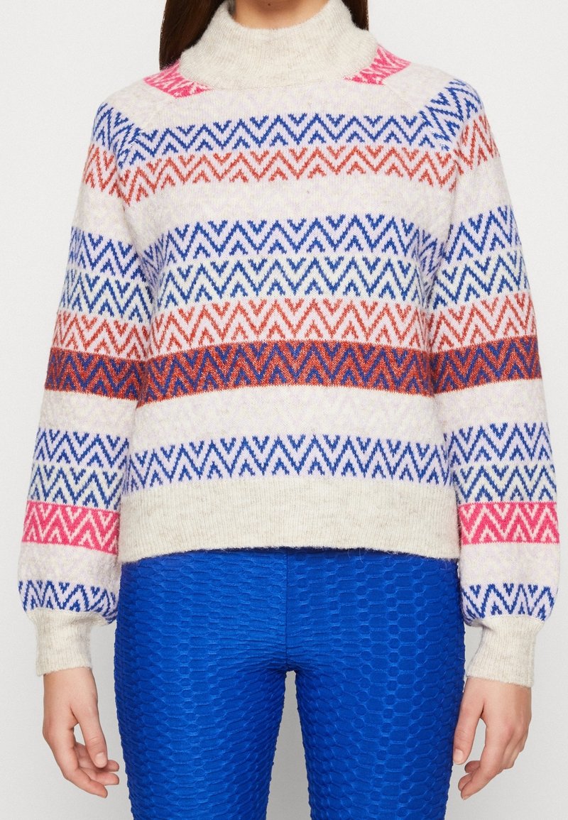 Personne portant un pull crème à rayures zigzag roses, rouges et bleues, associé à un pantalon bleu vif texturé, les mains détendues le long du corps.