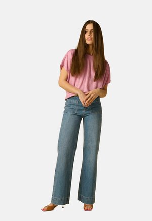 Vrouw met lang bruin haar die een loszittend roze T-shirt draagt, hoog getailleerde, wijde blauwe jeans en roze sandalen met open tenen, staand met gevouwen handen.