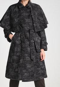 Černobílý vzorovaný vlněný kabát s návrhem pláště, páskem v pase a knoflíkovými detaily. Dlouhé rukávy s knoflíkovými detaily na manžetách.