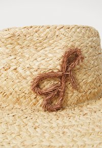 Pălărie din paie cu un design împletit, având un accent decorativ maro care seamănă cu un nod sau cu un fundiță. Finisaj natural, texturat, de culoare bej deschis.