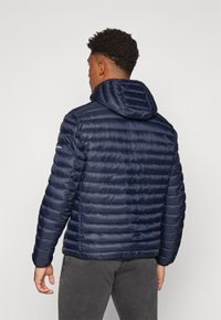 Tumši zils pūkains jakas ar kapuci, ar quilted sekcijām, gludu tekstūru un elastīgiem aprocēm. Aizmugures skats parāda regulējamu apkakles detaļu.