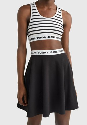Kobieta ubrana w czarną spódnicę i czarno-biały paskowany krótki top, oba z białymi elastycznymi taśmami z napisem "TOMMY JEANS."