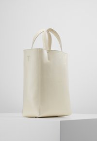 Krämfärgad läder tote-väska med dubbla handtag, stående upprätt på en vit pelare mot en slät ljusbakgrund.