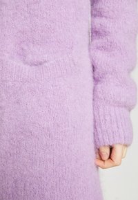 Gros plan sur une main près du poignet d'un pull tricoté doux lavande avec une poche texturée visible sur le vêtement.