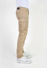 Zijaanzicht van een persoon in beige cargobroeken en witte sneakers, staande tegen een effen witte achtergrond.