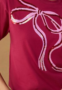 Fuchsia t-shirt med en stor rosett-design gjord av rosa och röda paljetter. Korta ärmar, jämn textur och avslappnad passform.