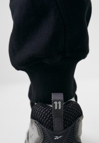 Schwarze Jogginghose über einem grauen Sportschuh, der ein strukturiertes Mesh-Obermaterial und einen Tab mit der Zahl "11" in Weiß hat.