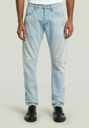 Mann trägt hellblaue Jeans und schwarze Lederschuhe und steht vor einem einfarbigen Hintergrund.