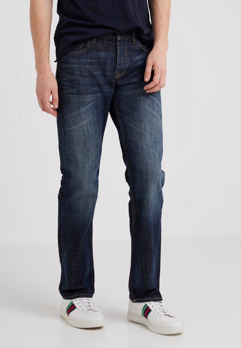 Tmavě modré džíny s rovnými nohavicemi, designem pěti kapes a jemným vyblednutím, doplněné bílými teniskami se multibarevnými pruhy.