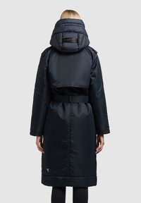 Temno modra podložena jakna z veliko kapuco, pasom s pasom, stranskimi žepi in logotipom na hrbtni strani. Gladek tekstura in dolžina do kolen.