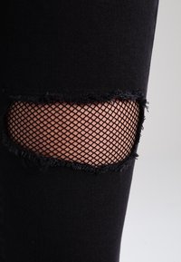 Jeans de mezclilla negros con una rodilla desgastada que expone un material de red negra debajo, junto con bordes deshilachados alrededor del desgarro.