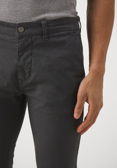 Pantalon en coton noir avec fermeture à bouton, devant plat et poches latérales. Le tissu semble lisse avec un léger éclat. Main reposant sur la jambe.