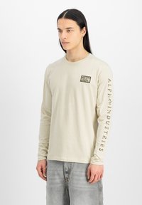 Camiseta de manga larga beige de algodón, con un pequeño parche en el pecho y "ALPHA INDUSTRIES" impreso a lo largo de la manga izquierda en un color contrastante.