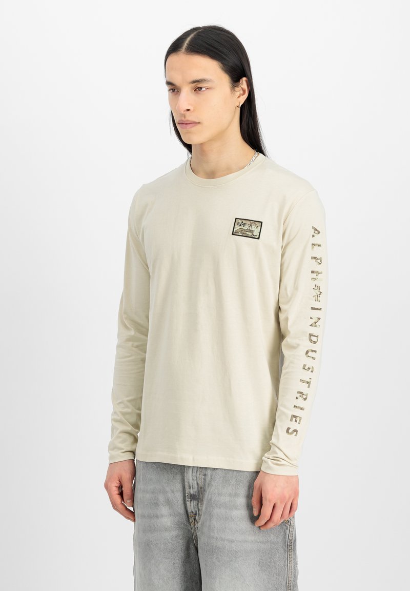 Camiseta de manga larga beige de algodón, con un pequeño parche en el pecho y "ALPHA INDUSTRIES" impreso a lo largo de la manga izquierda en un color contrastante.