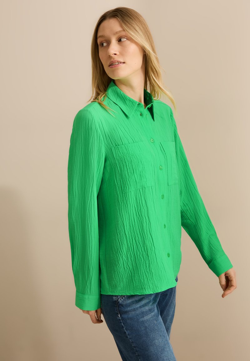 Camicia verde a maniche lunghe con texture, design a bottoni, dotata di tasca e colletto a punta, abbinata a jeans in denim blu.