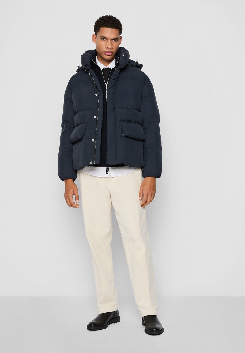 Veste matelassée marine avec capuche amovible, texture matelassée, boutons-pression à l'avant et quatre grandes poches à l'avant, portée avec un pantalon clair et des bottes noires.