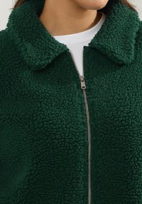 Zielona polarowa kurtka z teksturowaną powierzchnią, kołnierzem i metalowym zamkiem błyskawicznym z przodu. Noszona na białej koszuli.