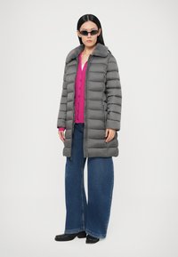 Naine kannab pikka halli sulejope karvaga kraega, roosat kardigani, laiade säärtega siniseid teksapükse, musti saapaid ja kitsaid musti päikeseprille.