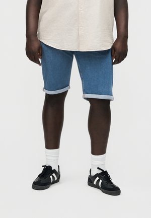 Osoba nosiaca vyhrnuté modré džínsy, biele ponožky a čierne tenisky s bielymi pruhmi, stojaca na pozadí s jednofarebnou stenou.