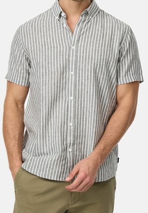 Mężczyzna ubrany w szarą i białą koszulę w pionowe paski z krótkim rękawem, zapinaną na guziki, oraz khaki spodnie, z ręką w kieszeni, na jednolitym tle.