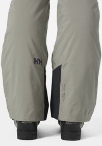 Pantalones impermeables grises con una textura suave, con la marca Helly Tech y detalles negros en los tobillos, combinados con botas negras que muestran un diseño de malla.