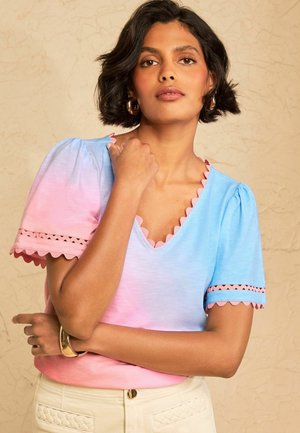Woman with short black hair wearing a pink and blue scalloped edge short-sleeve top and beige pants against a beige textured background.