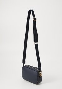 Kompaktna mornarsko plava crossbody torba sa zlatnim zatvaračem, teksturiranim trakama, pravokutnim oblikom i suptilnim logotipom na dnu.